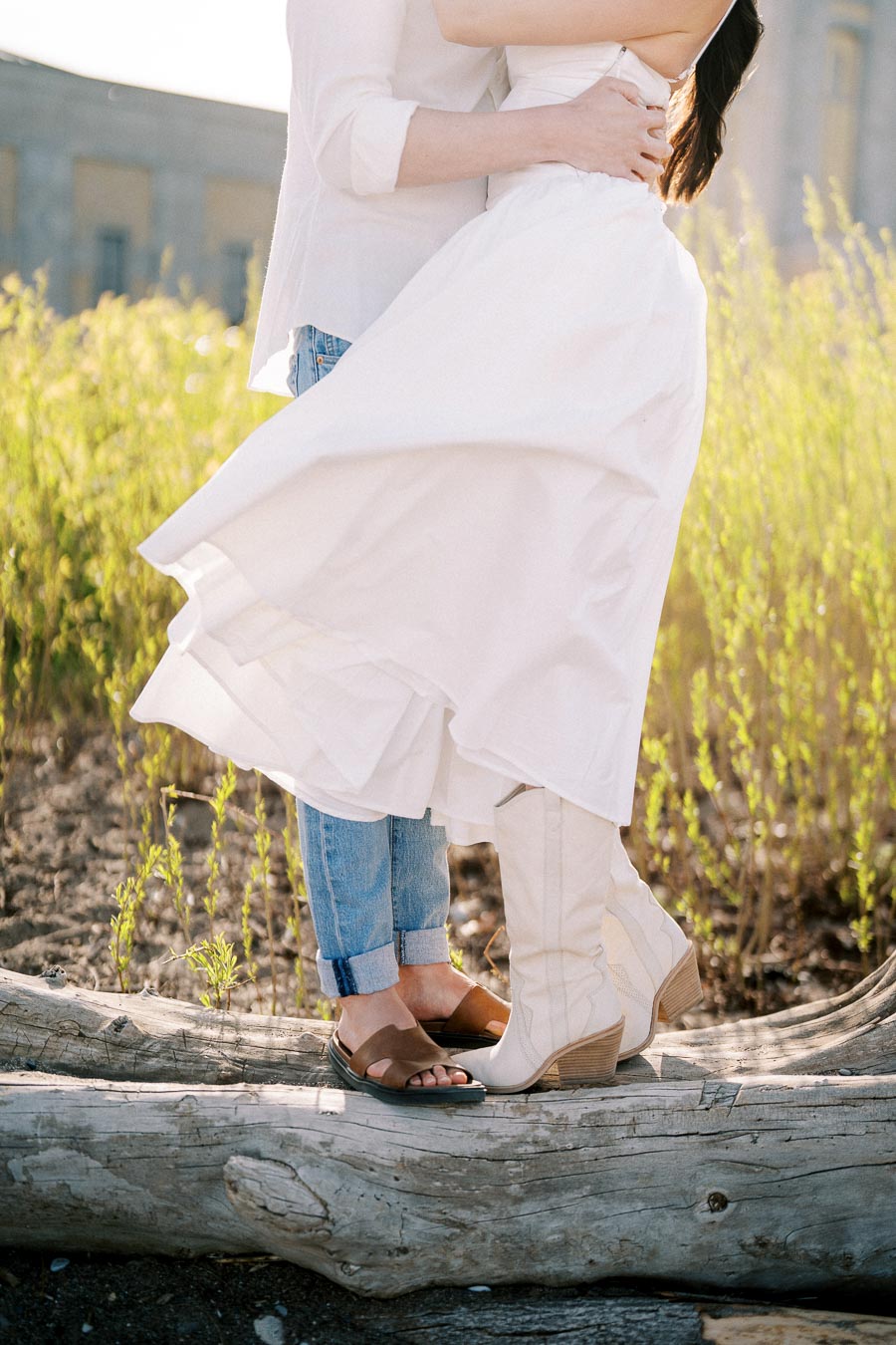 Couple embracing outdoors on a sunny day, woman in white dress and boots, standing on a log surrounded by greenery.