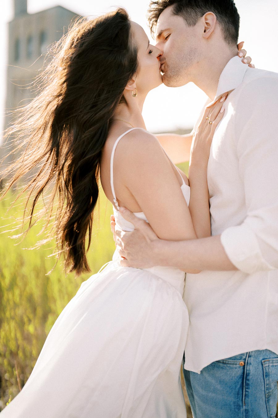 Couple sharing a romantic kiss in a sunlit field, with the woman in a white dress and the man in a white shirt and jeans.