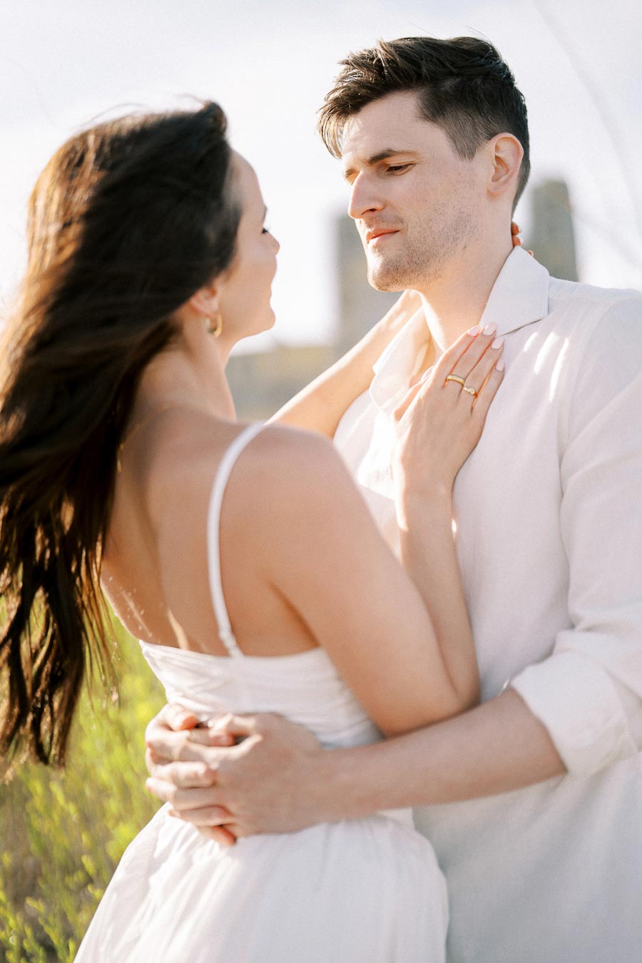 A romantic couple embraces outdoors, both dressed in white, with a serene, sunlit backdrop, depicting warmth and affection.