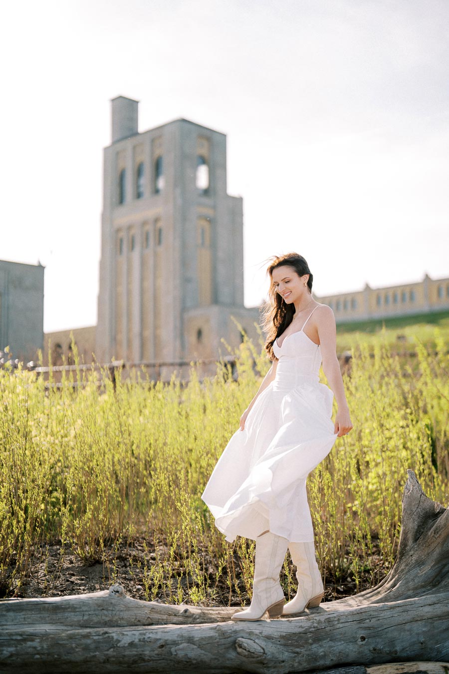 Woman in a flowing white dress standing on a log, surrounded by tall grass with a historic building in the background. Bright, sunny day with a natural, rustic setting.