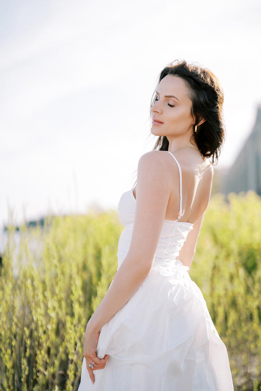 Woman in white dress standing in a serene outdoor setting with greenery, enjoying a sunny day.