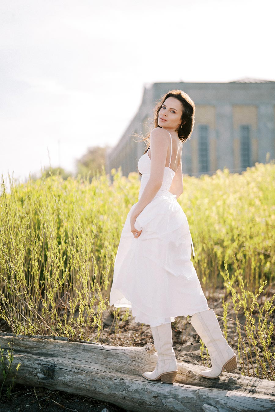 A woman in a flowing white dress and white boots stands on a log in a sunlit grassy field, with a stone building in the blurred background.