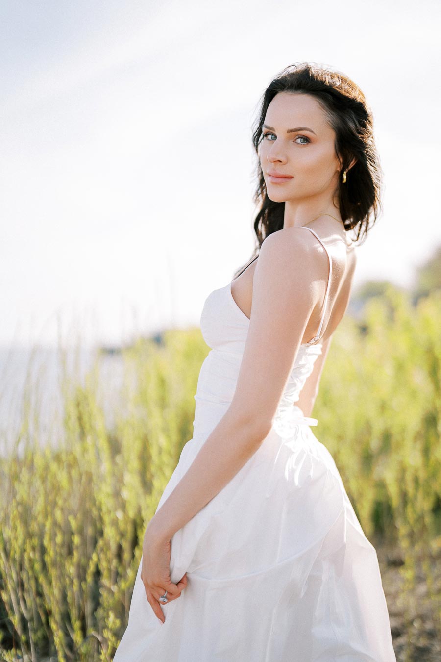 Young woman in a white dress standing in a sunny outdoor field, with green plants in the background