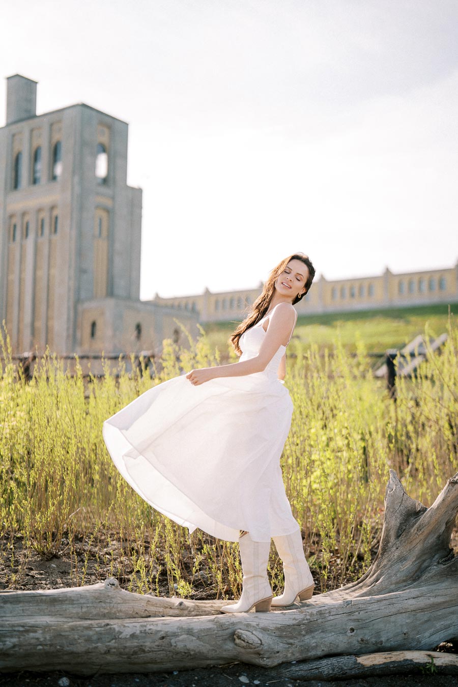 Woman in a flowing white dress standing on a log with tall grasses around, smiling with closed eyes, historical building in the background.