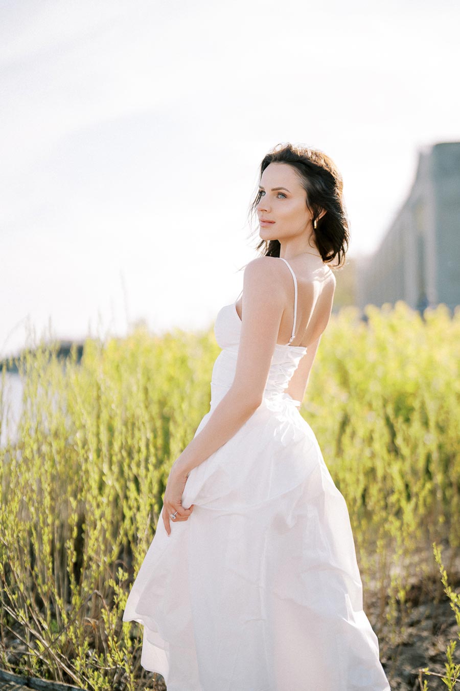 A woman in a white dress stands in a field of yellow flowers on a sunny day, with a serene expression and sunlight illuminating her hair.
