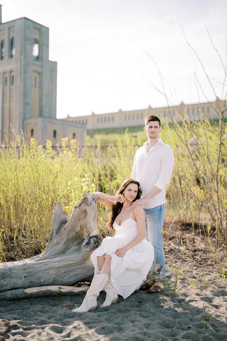 A couple in light clothing posed outdoors on sandy terrain with a stone tower in the background, surrounded by greenery and a large piece of driftwood.
