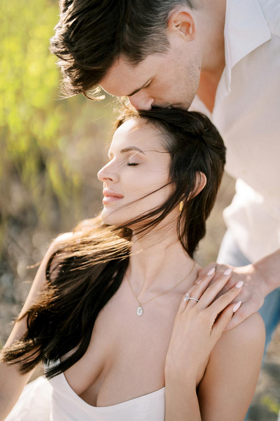 A romantic moment captured between a couple outdoors, with a man gently kissing the woman's forehead as she closes her eyes and smiles slightly, her hair flowing in the breeze. The sunlit background creates a serene and intimate atmosphere.