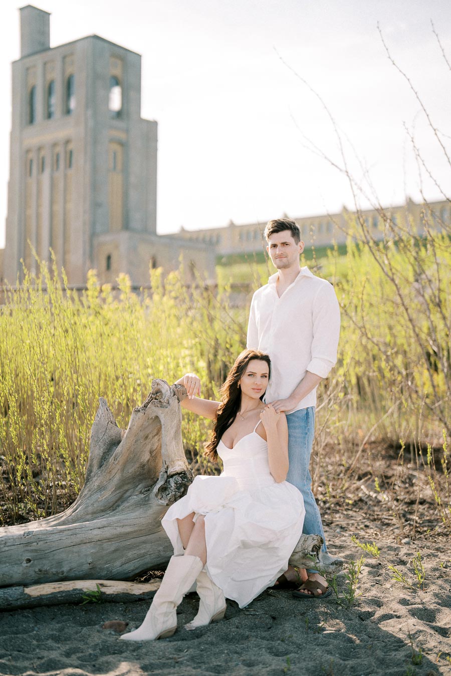 Young couple in casual and white attire posing outdoors near a large log, with an elegant historical building in the background and natural greenery surrounding them.