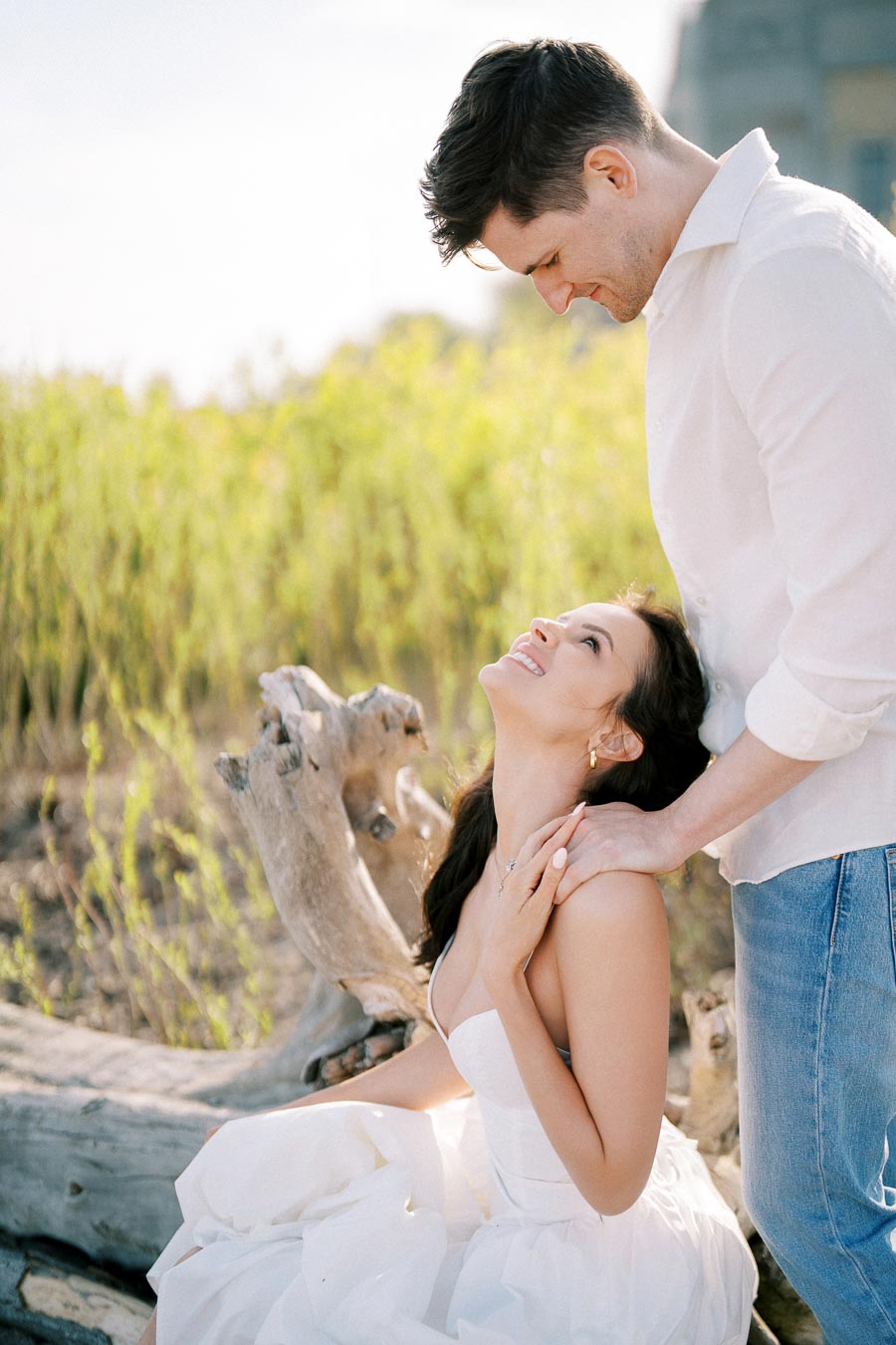 A couple embracing in a serene outdoor setting, with a woman in a white dress sitting on a log and looking up, smiling at a man in a white shirt and jeans, surrounded by greenery.
