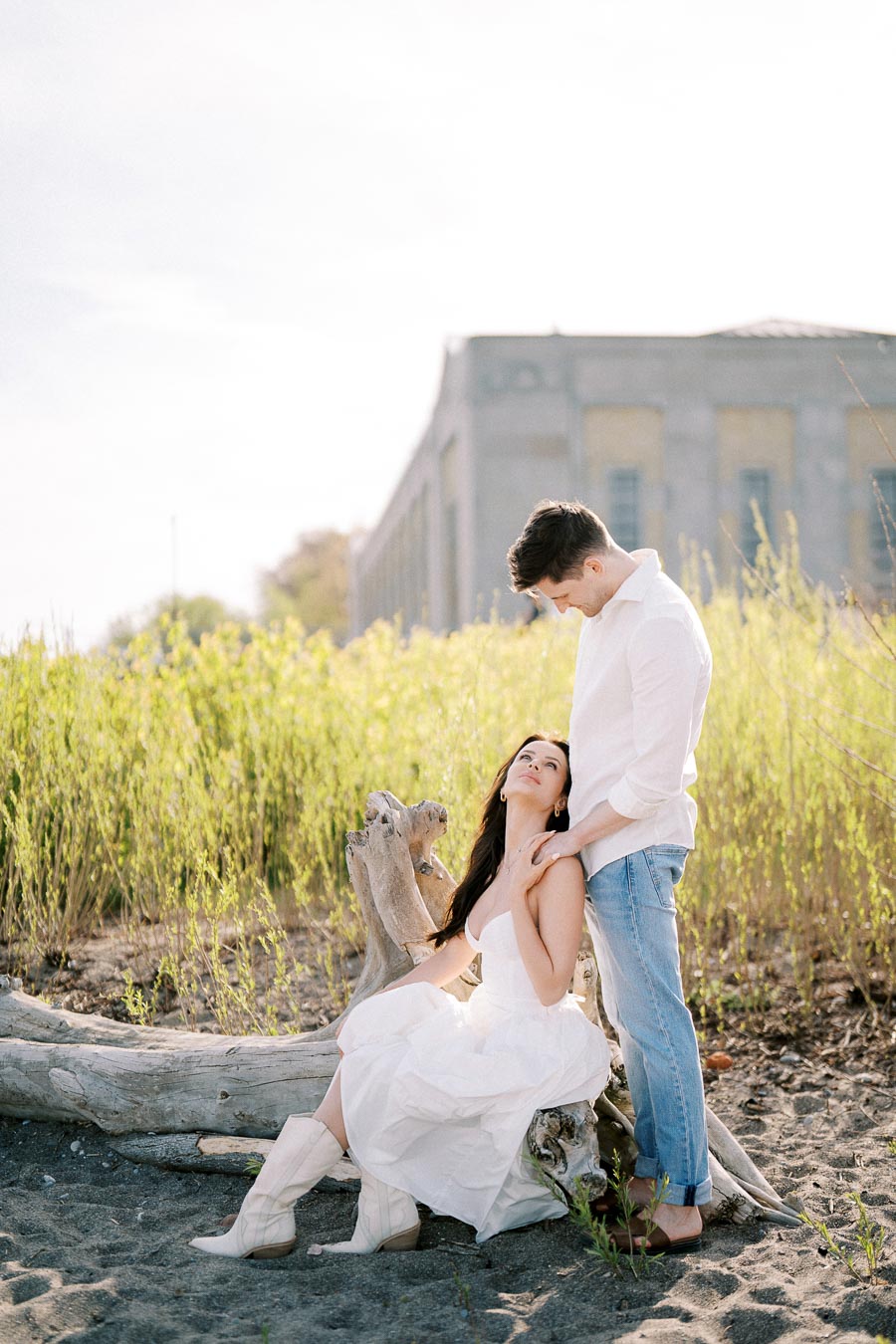A couple in casual attire shares an intimate moment on a sandy beach. The woman, wearing a white dress and boots, sits on a driftwood log while gazing up at the man in a white shirt and jeans. A historic building and lush greenery can be seen in the background, evoking a romantic outdoor setting.