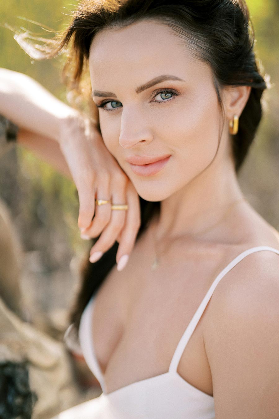 Young woman with brown hair poses in a sunny outdoor setting, wearing a white camisole and gold earrings.