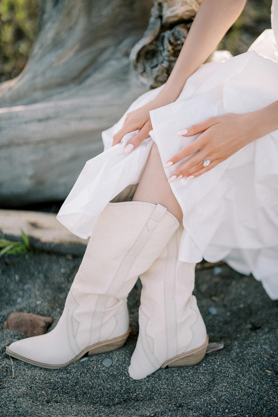 Bride in a white dress and cowboy boots sitting on sand, showcasing elegant manicure and engagement ring, highlighting rustic bridal fashion.