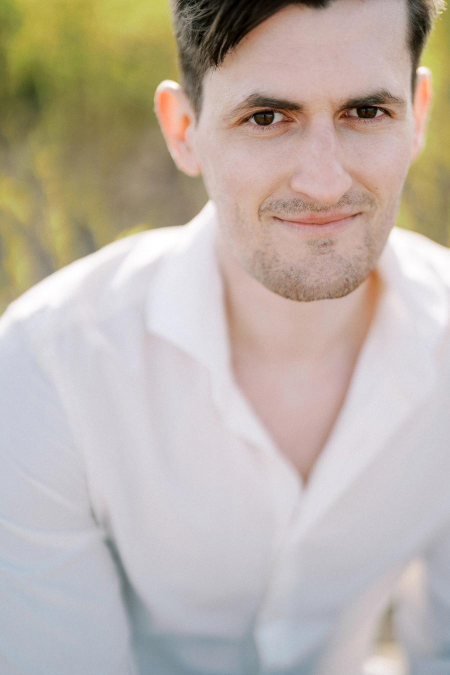 Portrait of a person smiling slightly, wearing a white shirt with a blurred natural background on a sunny day.