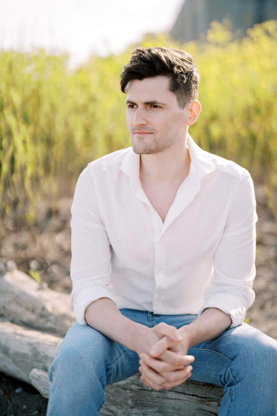 A man in a white shirt and jeans sitting on a log outdoors, looking thoughtful with green foliage in the background, under natural sunlight.