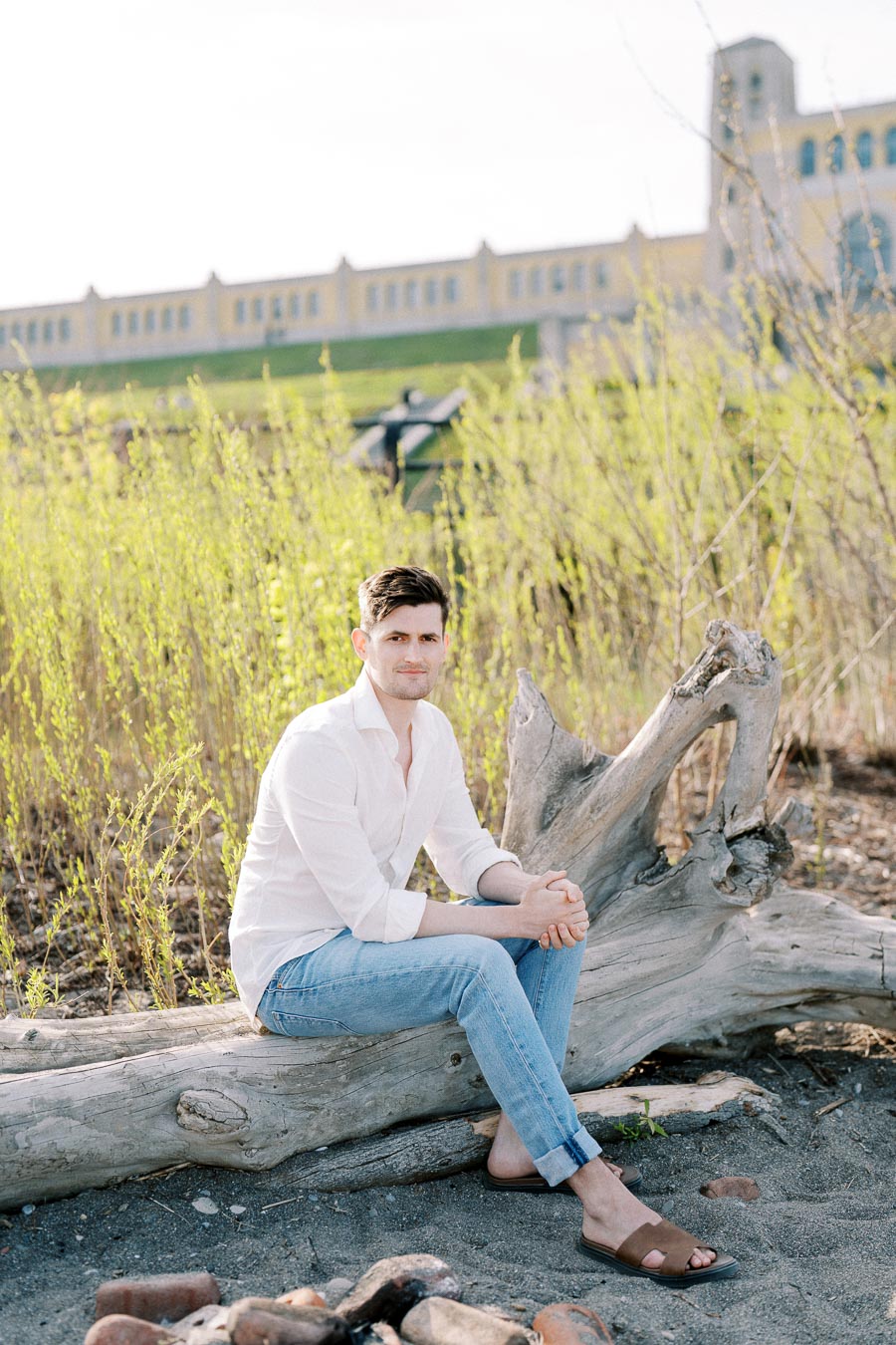 A person in a white shirt and jeans sits on a large piece of driftwood with green foliage and a building in the background on a sunny day.