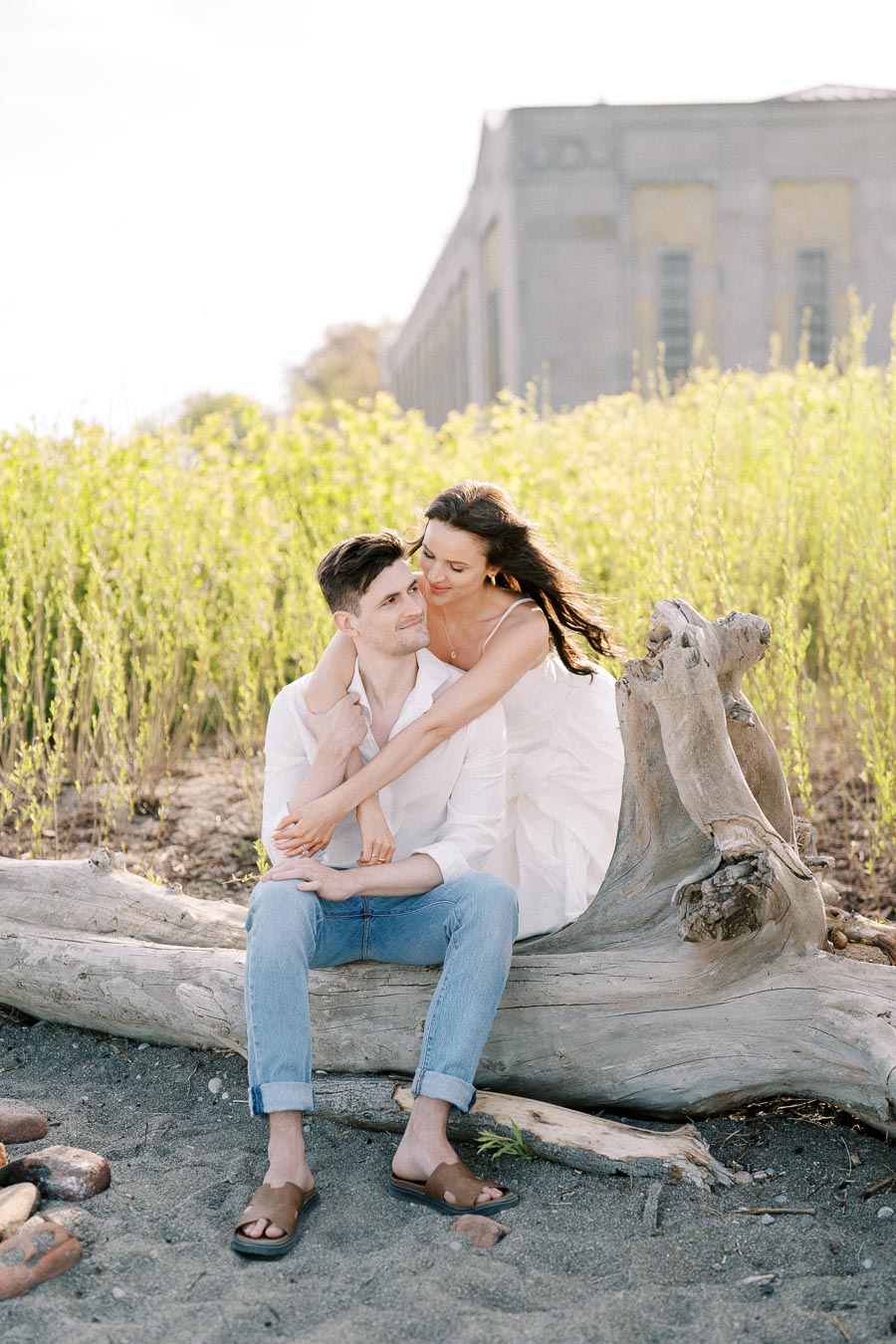 A couple embracing on a beach sitting on a large driftwood log, with greenery and an industrial building in the background, under bright daylight.