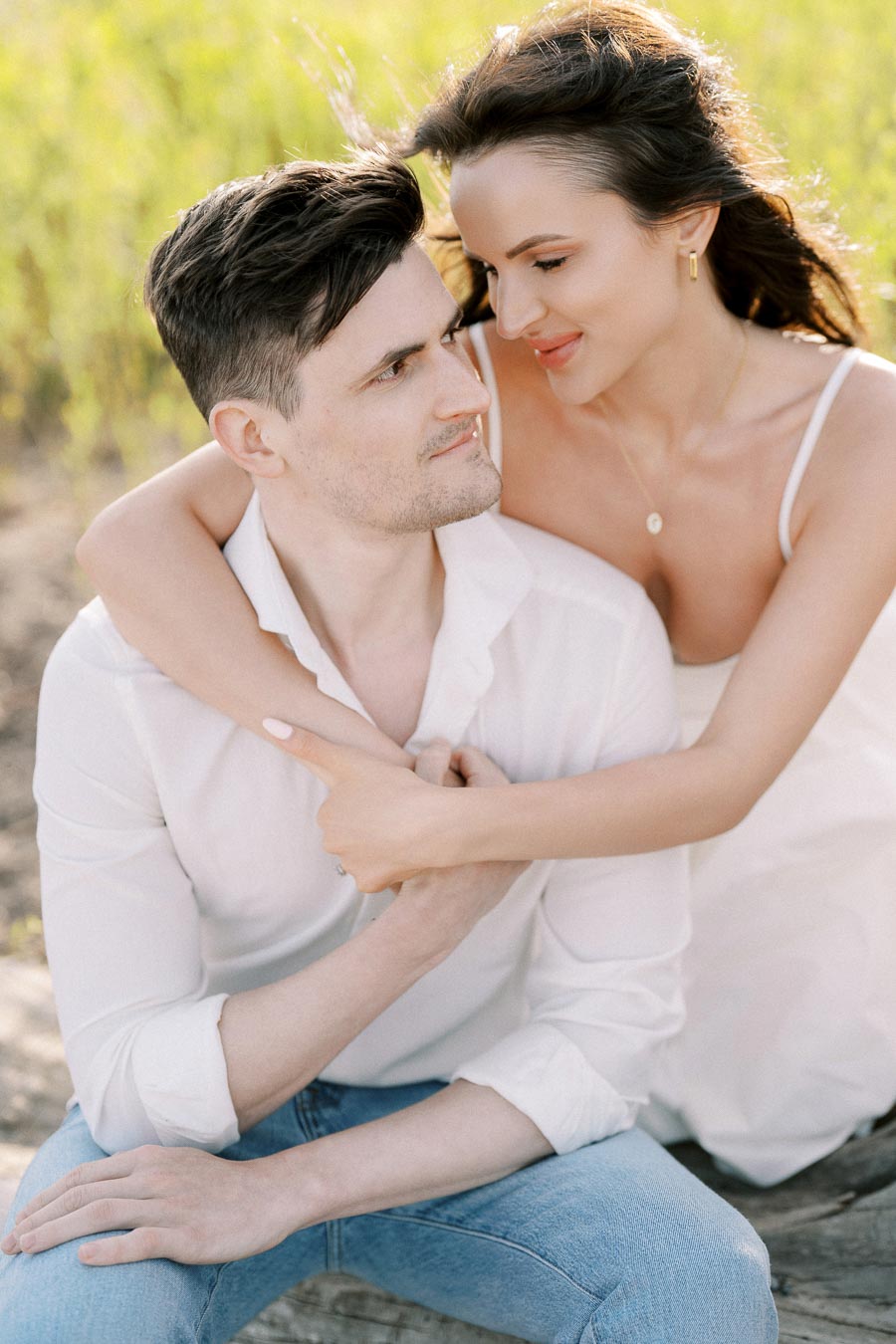 A romantic couple embracing outdoors with a man in a white shirt and jeans sitting, while a woman in a white dress wraps her arms around him, surrounded by a green field.