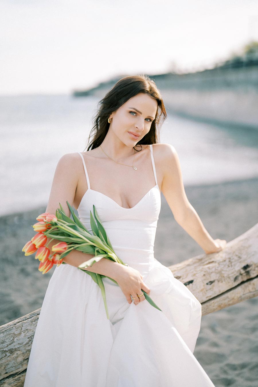 A woman in a white dress holding a bouquet of orange and yellow tulips poses by a beachside log, with the ocean and a clear sky in the background.