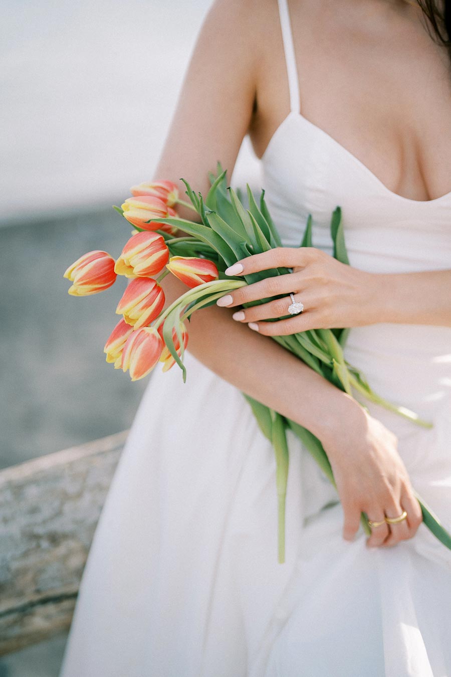 Bride in white dress holding a bouquet of orange tulips, showcasing an elegant engagement ring on her finger.