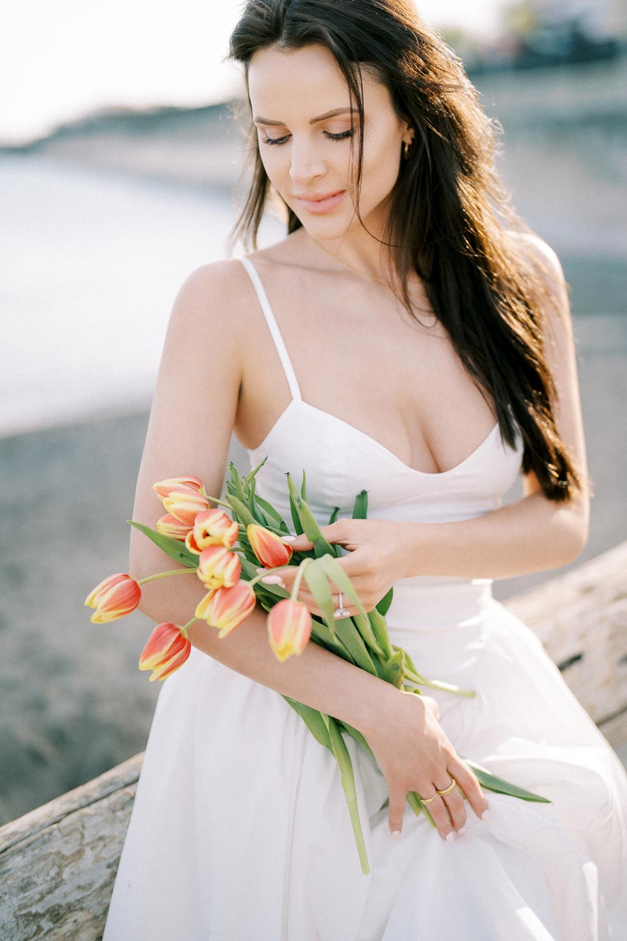 A woman in a white dress holding a bouquet of orange and yellow tulips on a beach background.
