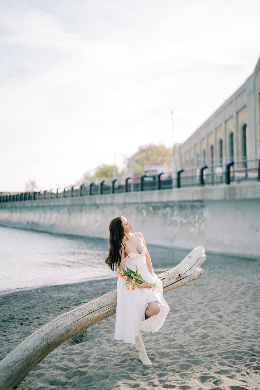 A woman in a white dress sits on a driftwood log by the waterfront, holding a bouquet of orange tulips. The scene captures a serene moment with a backdrop of a river and industrial architecture on a sunny day.