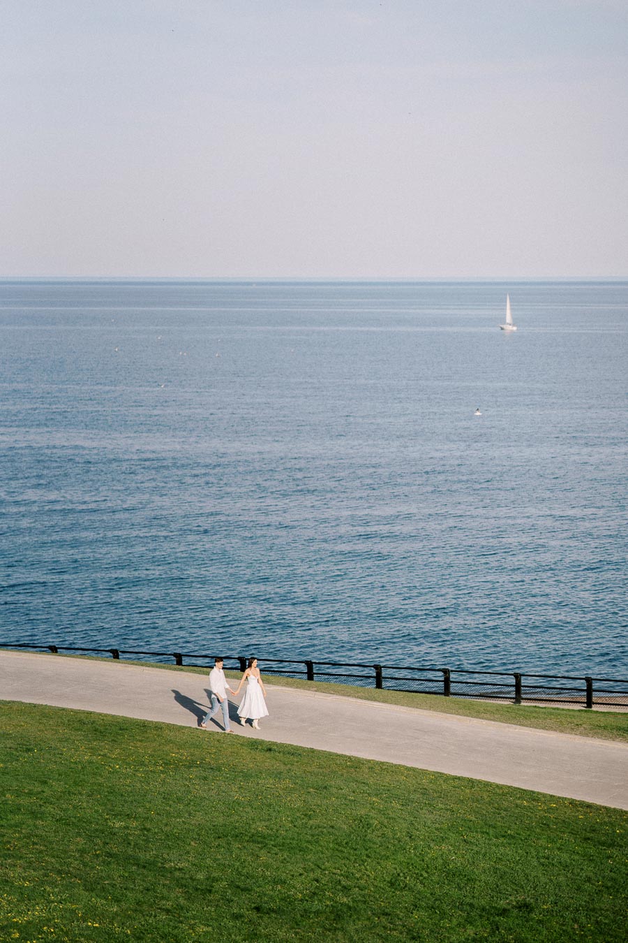 A couple holding hands walks along a scenic coastal path with a vast blue ocean and sailboat in the background, under a clear sky.
