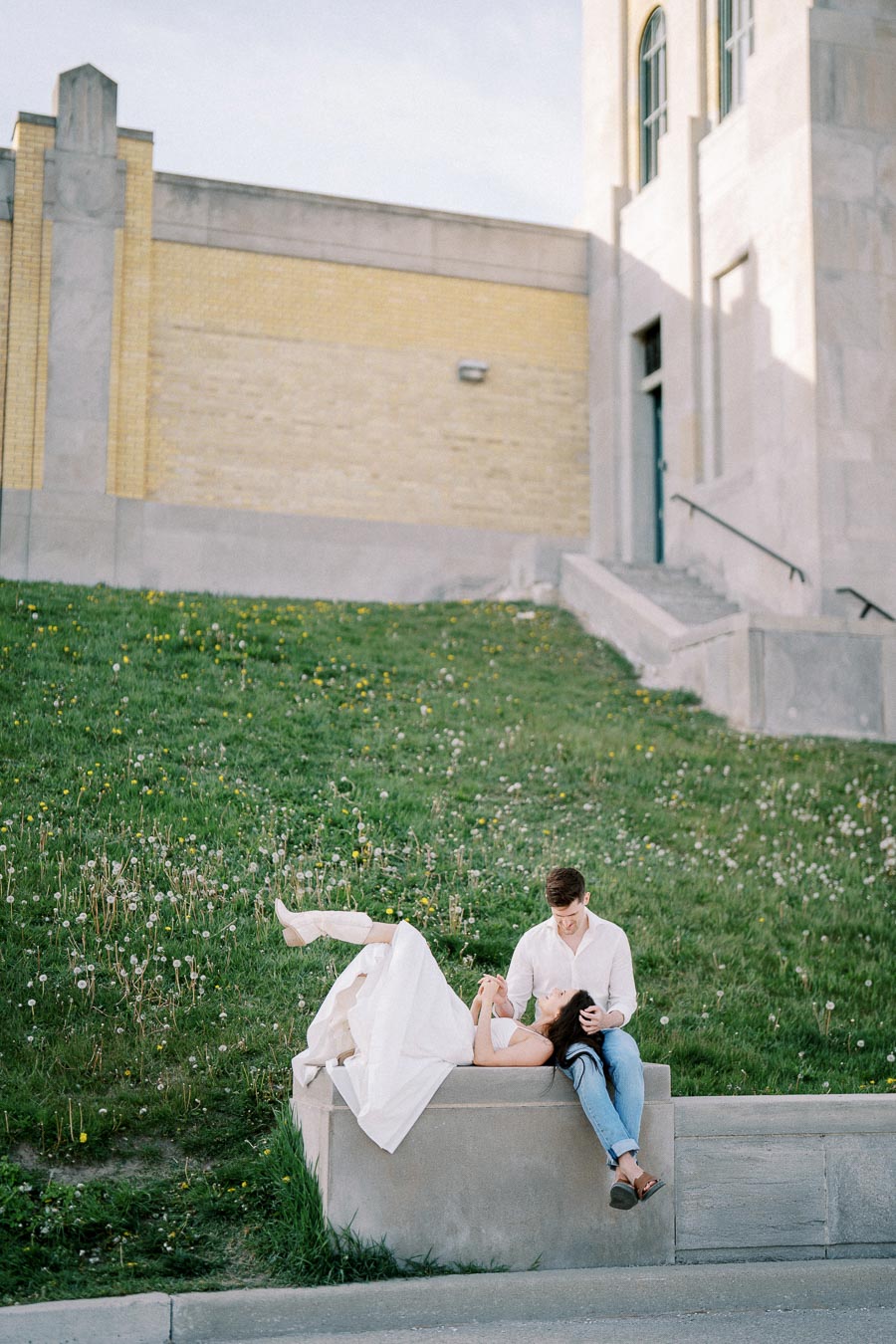 A couple enjoying a romantic moment on a grassy hillside, with the woman lying back on a concrete bench and the man sitting beside her on a sunny day, near a modern building.