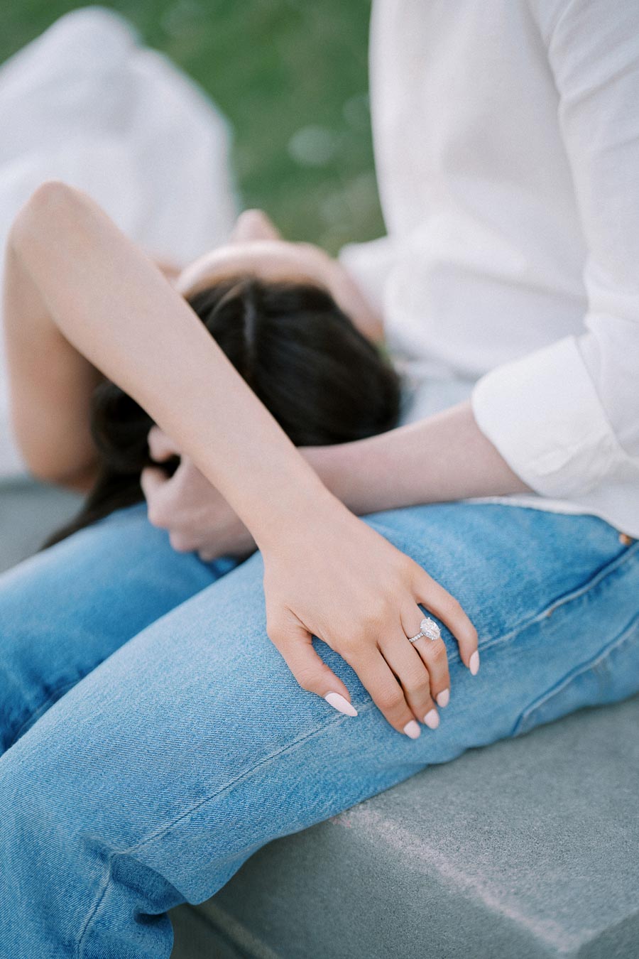 A woman with an elegant engagement ring rests her head on the lap of a person wearing blue jeans and a white shirt, symbolizing love and intimacy.