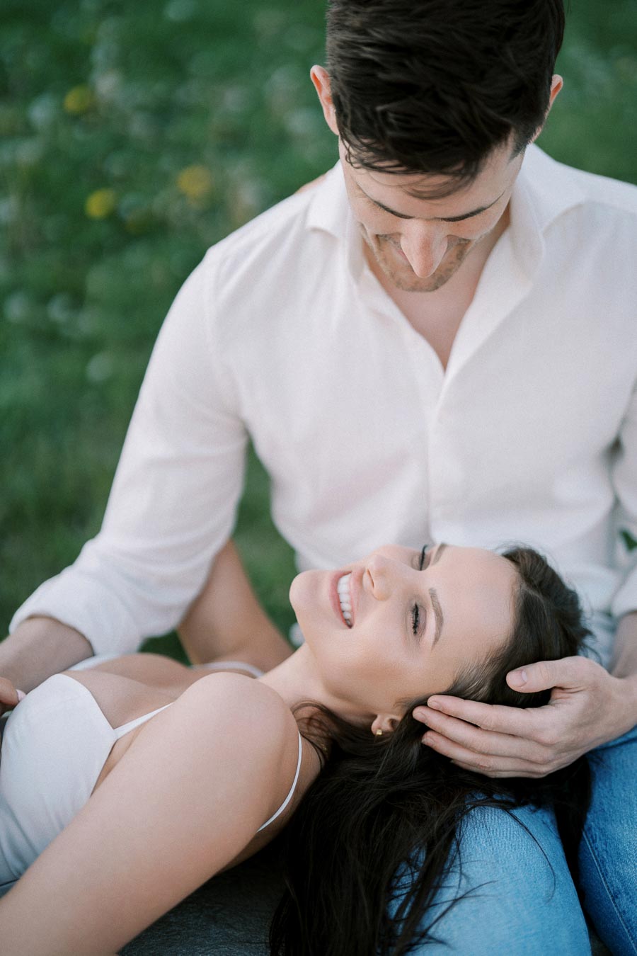 A couple enjoying a romantic moment outdoors; a woman lying on her partner's lap, smiling and relaxed, surrounded by a grassy garden setting.