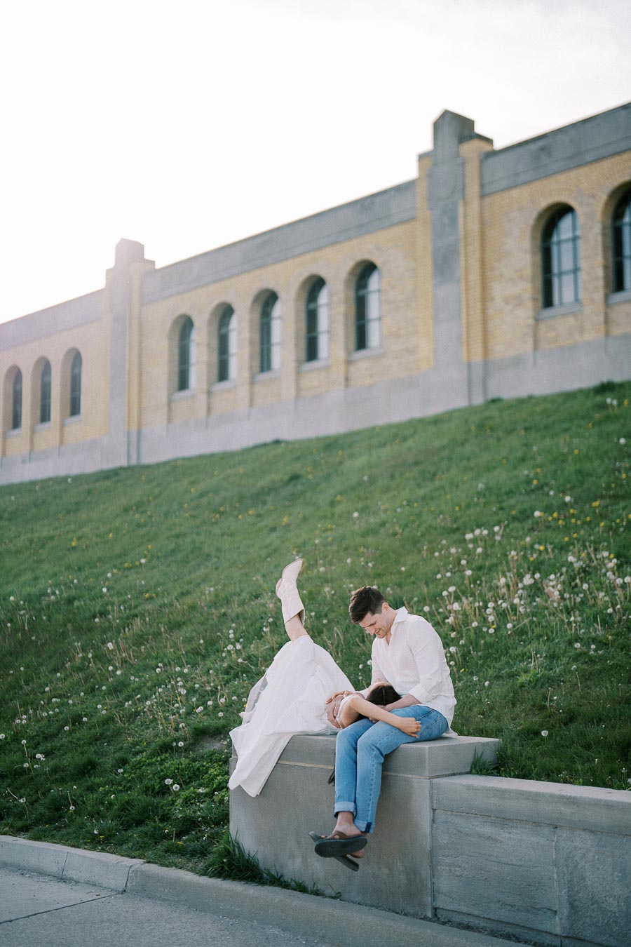 A couple in casual attire enjoys a playful moment outdoors on a grassy hill by a historic building, with a woman playfully leaning back over a man's lap, both smiling.