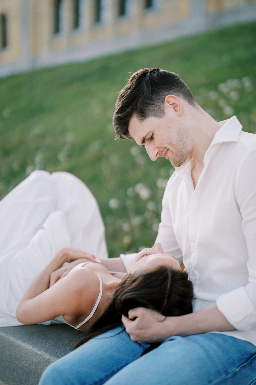 A couple sitting outdoors on a grassy hill, with the woman lying in the man's lap, both wearing white clothes and embracing affectionately.