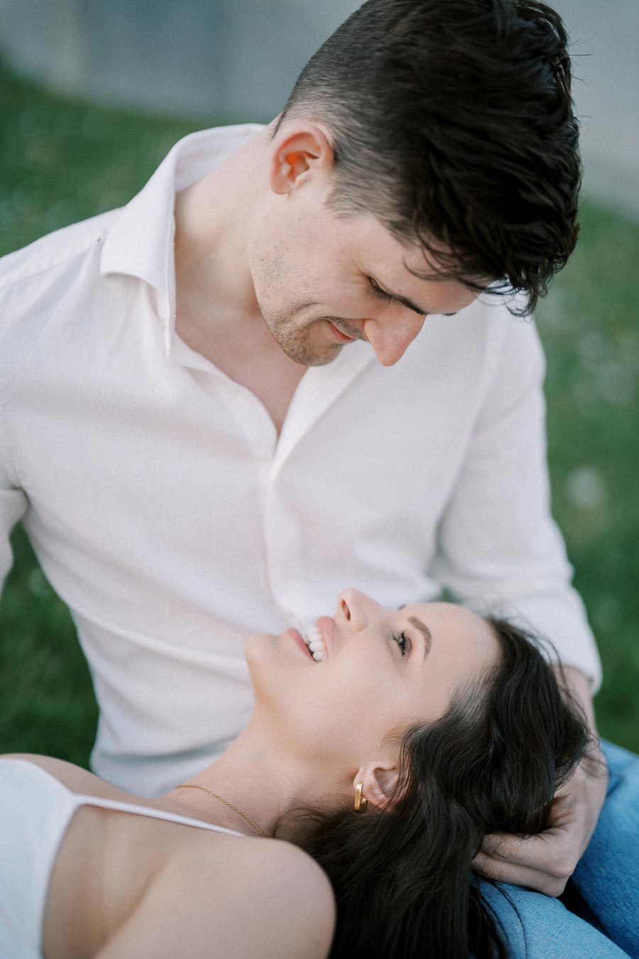 Romantic couple enjoying a serene moment in a park, with a man in a white shirt looking down lovingly at a smiling woman resting her head on his lap.