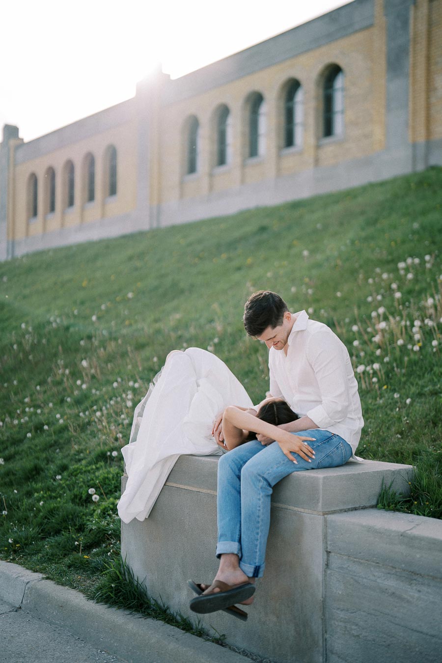 Romantic couple sitting on a stone ledge, with the woman resting her head on the man's lap, set against the backdrop of an old brick building and a grassy hill, capturing a serene outdoor moment.