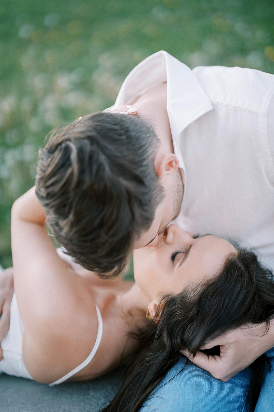 A romantic couple sharing an intimate moment while kissing outdoors, dressed in casual summer clothing, surrounded by a blurred green background.