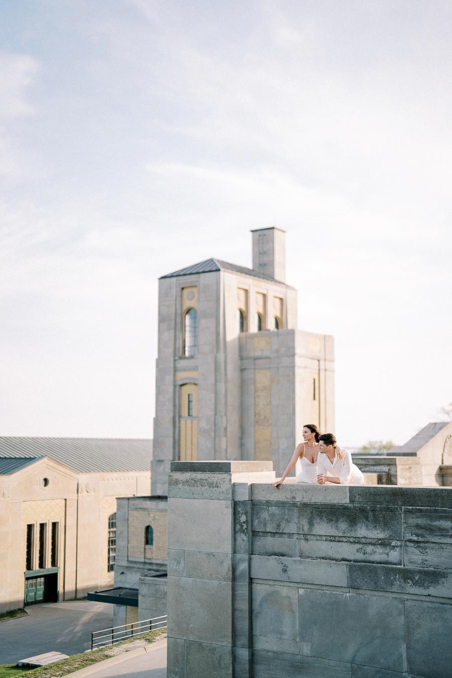 Two people in white attire lean against a concrete ledge, with a large, historic stone building in the background under a clear blue sky.