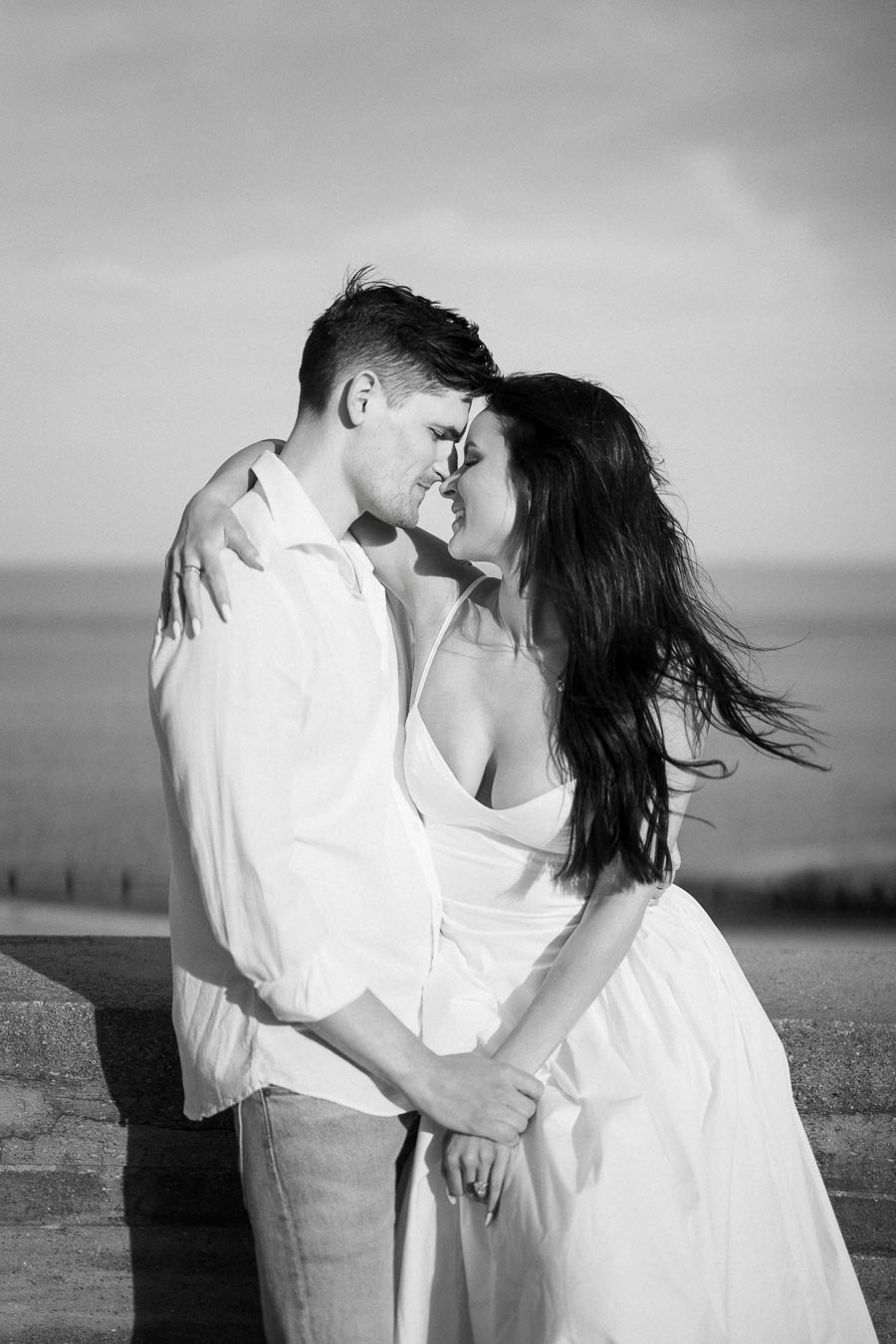 A black and white photo of a couple embracing intimately, standing on a beach with the ocean in the background, both wearing casual white clothing.