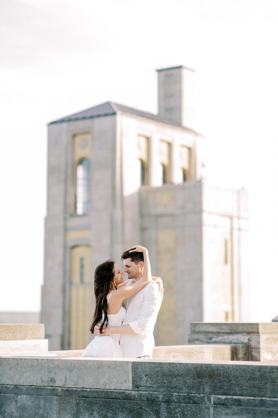 A couple embracing on a rooftop with a historic stone building in the background, dressed in light-colored attire, capturing a romantic moment.