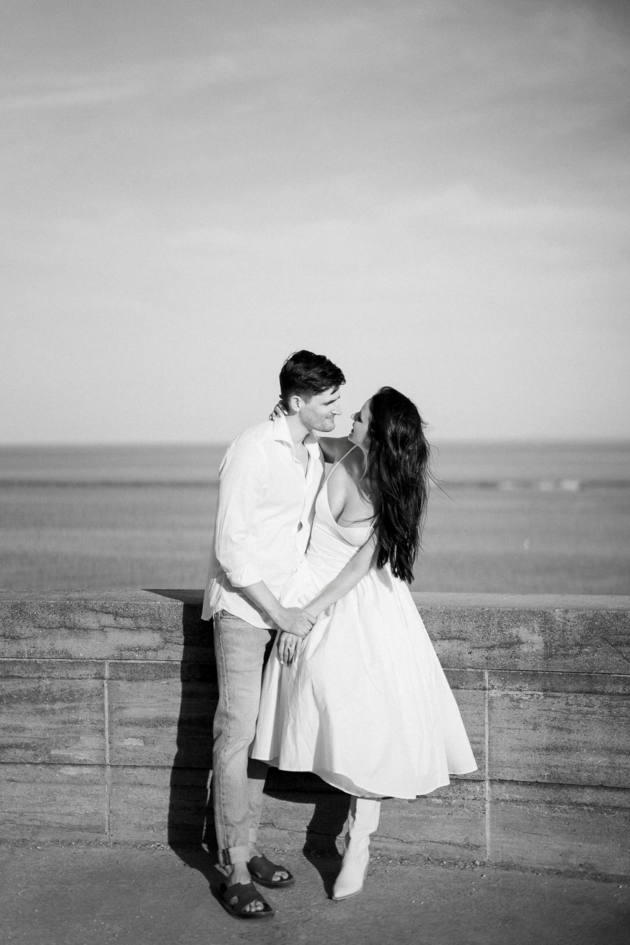 A couple shares an intimate moment overlooking the ocean, dressed in casual summer attire, capturing a romantic scene by the seaside stone wall in black and white.