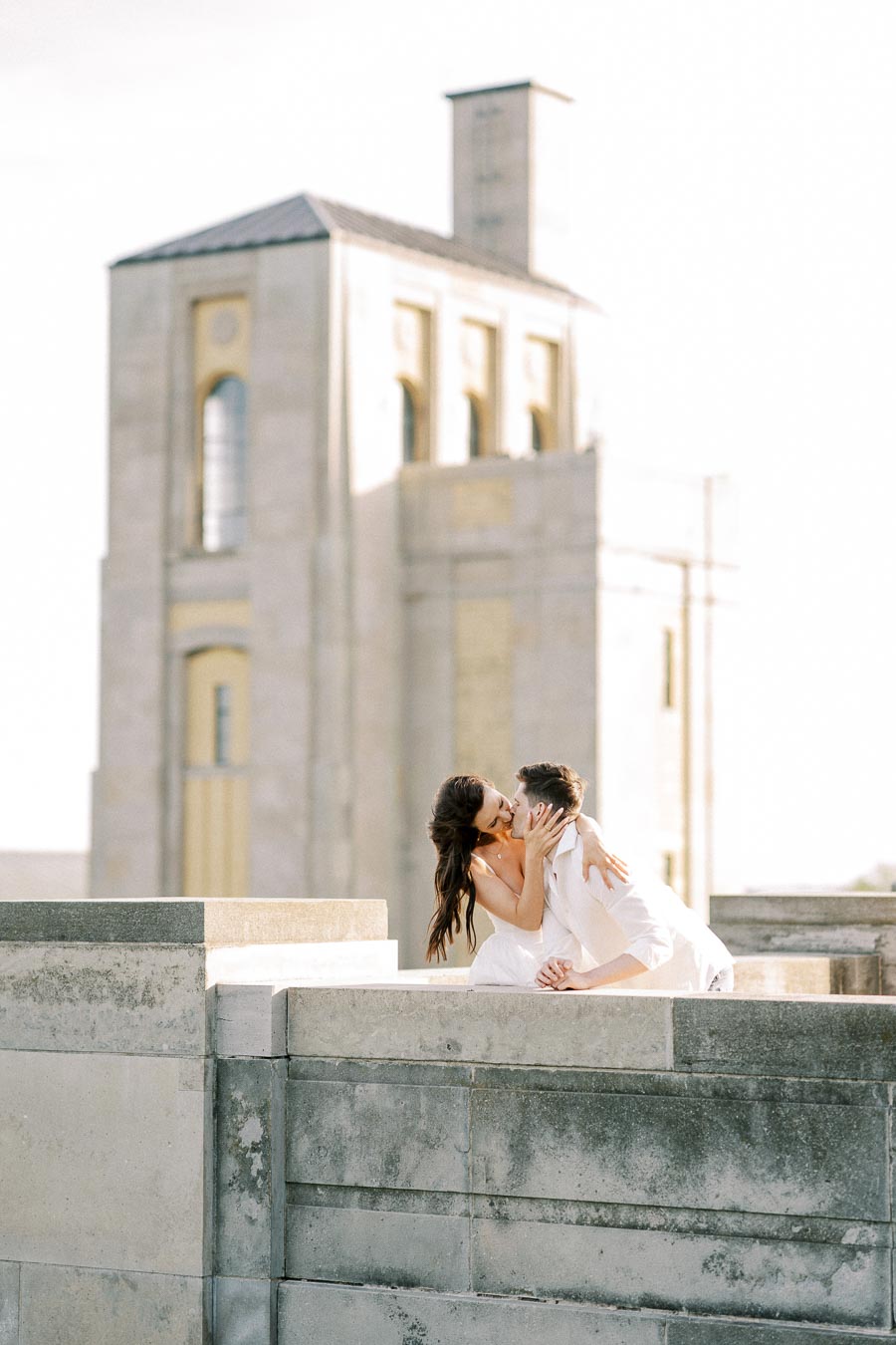 Romantic couple sharing a kiss on a rooftop with a historic building in the background under soft sunlight.
