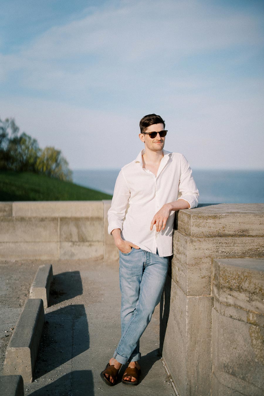 Man wearing sunglasses, white shirt, and jeans, leaning against a stone railing near a waterfront on a sunny day.