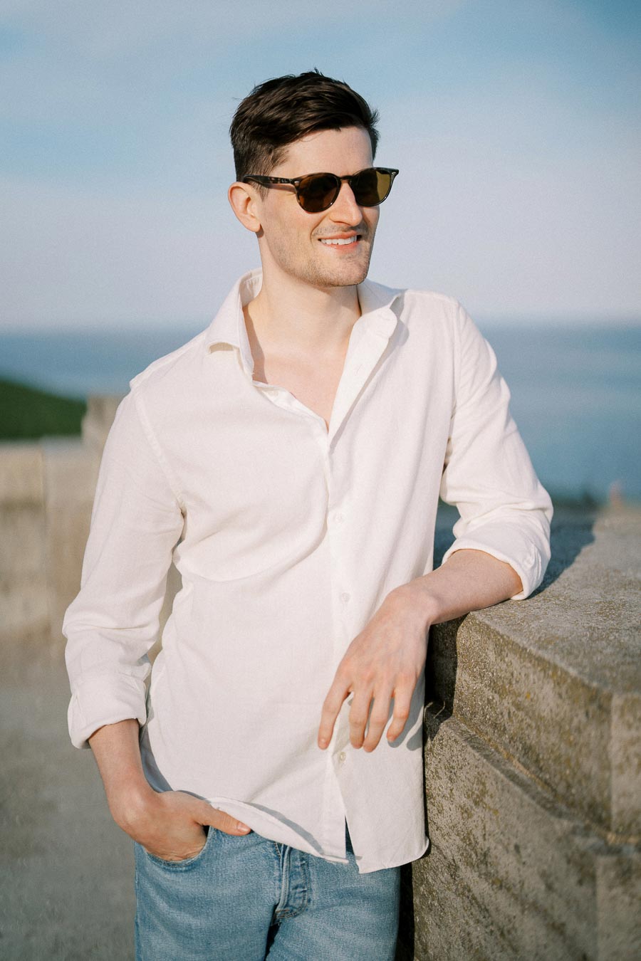 A person in sunglasses wearing a white shirt and jeans stands by a stone wall, smiling under a clear blue sky.