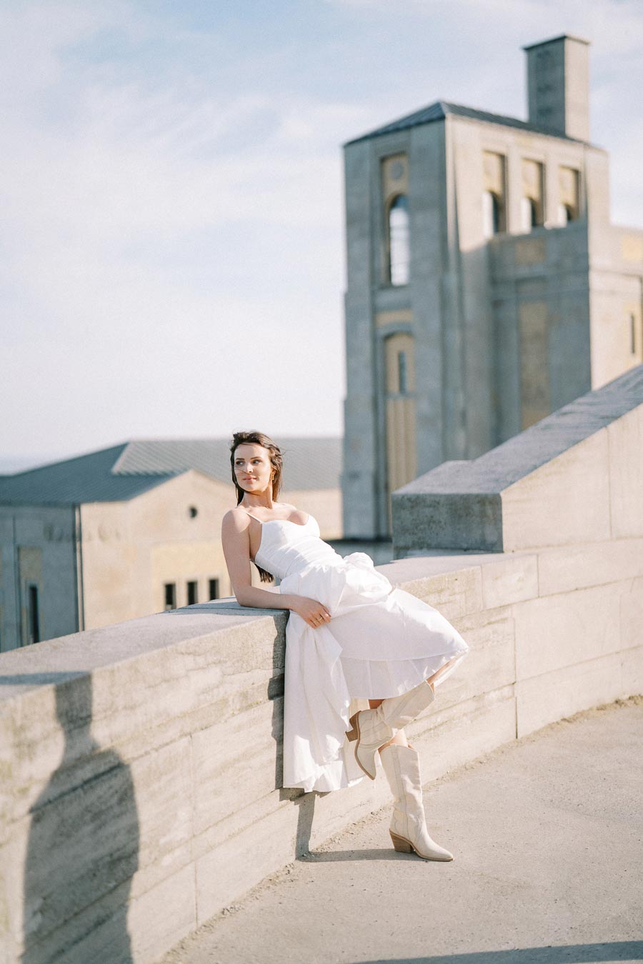 Young woman in a white dress and boots leaning against a stone wall with a historic building in the background, enjoying a sunny day.