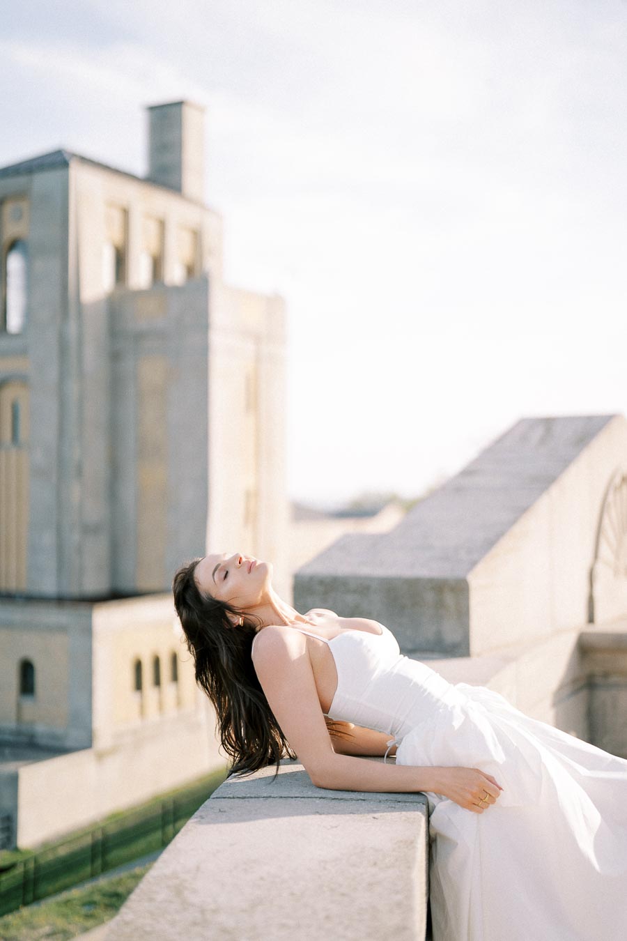 A woman in a white dress leans back against a stone railing, with an architectural building in the background under a clear sky, creating a serene and elegant scene.