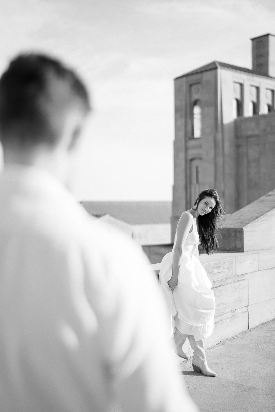 Black and white photo of a woman in a flowing white dress leaning against a stone ledge, with a blurred figure in the foreground and a historic building in the background, capturing a serene and elegant urban scene.
