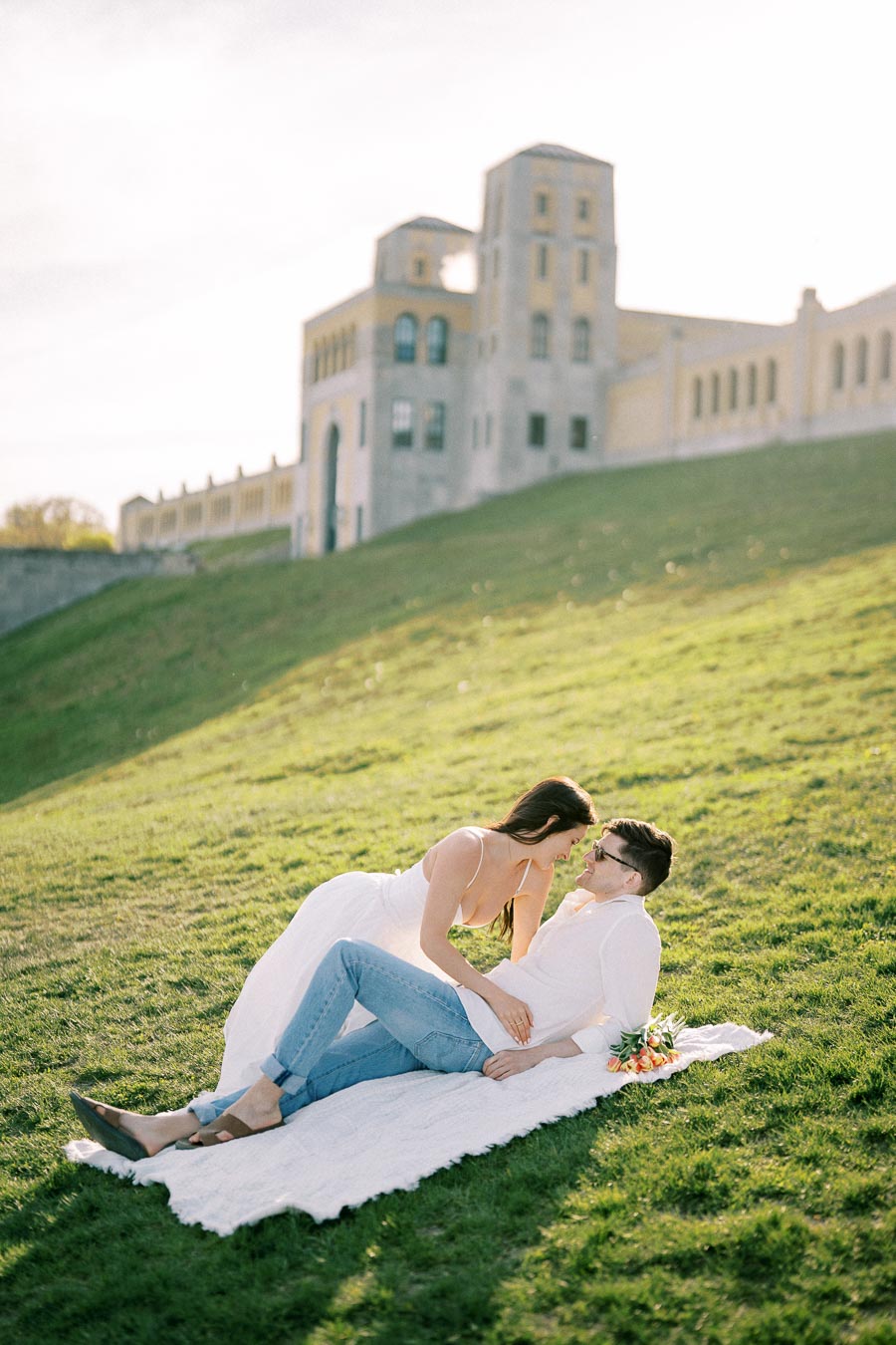 A couple embracing on a picnic blanket in a grassy area in front of a historic building on a sunny day.