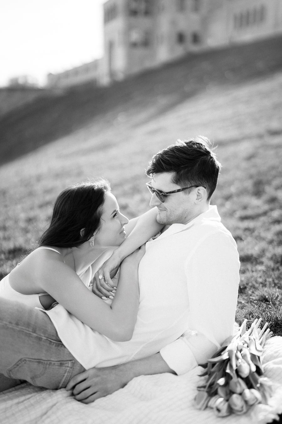 Romantic black and white photo of a couple embracing on a grassy hillside, with a bouquet of tulips nearby, capturing a serene and intimate moment.