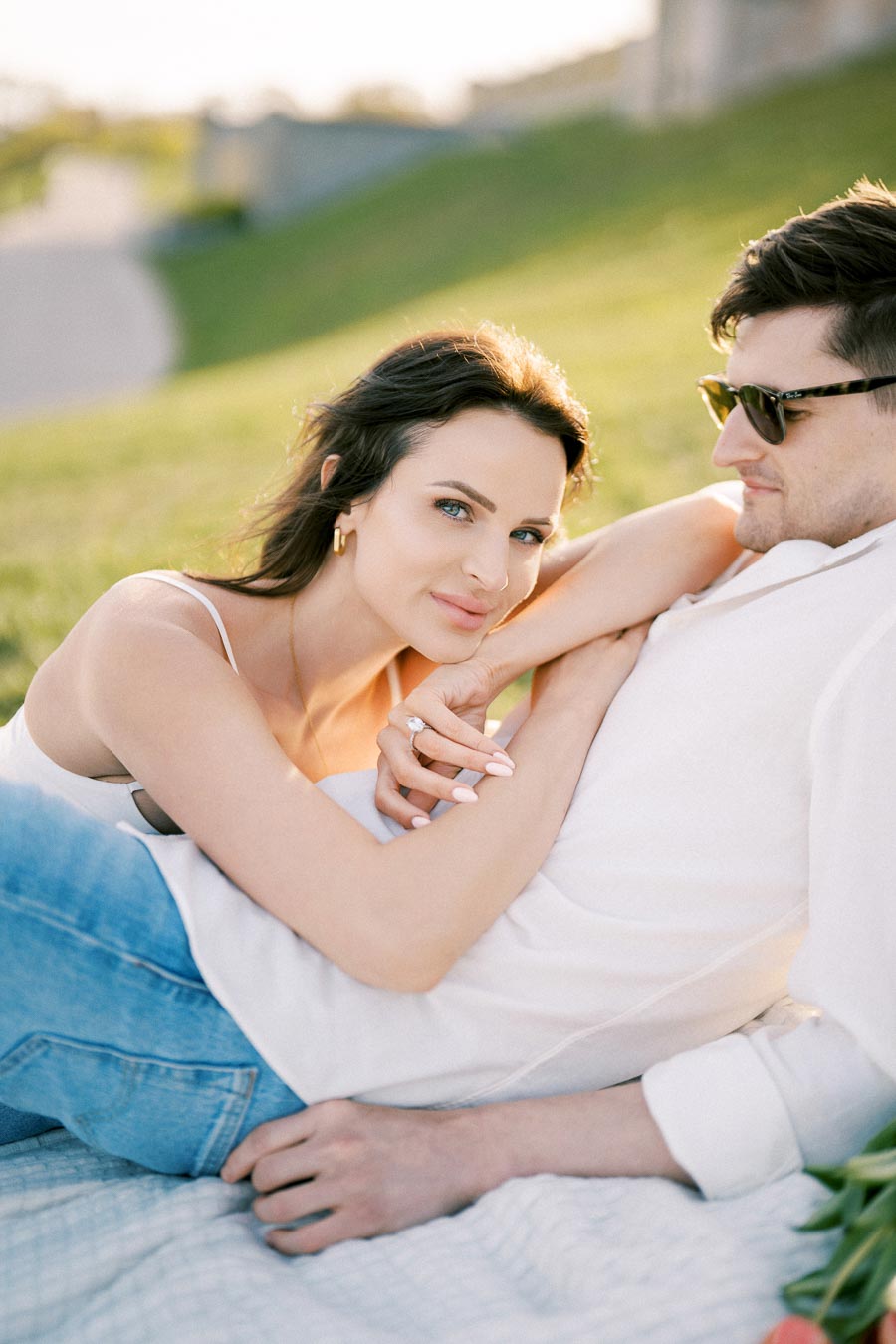 A couple enjoying a sunny day outdoors, lounging on a picnic blanket in a lush green park. The woman, wearing a white tank top, gazes at the camera while leaning against her partner, who sports sunglasses and a white shirt. The setting suggests a relaxing, romantic atmosphere.