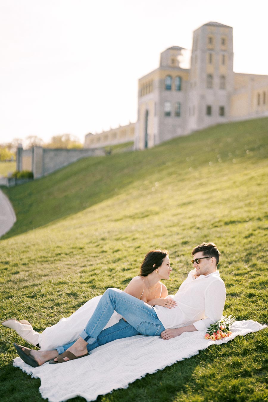 A couple relaxing on a picnic blanket in a sunlit park, with a historic stone building in the background. The woman is looking at the man lovingly while holding a bouquet of tulips, creating a romantic outdoor scene.