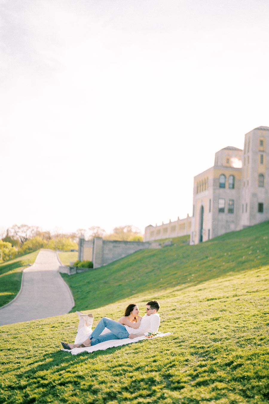 A couple enjoying a relaxing picnic on a grassy hill with a historic building in the background, under a clear sky.