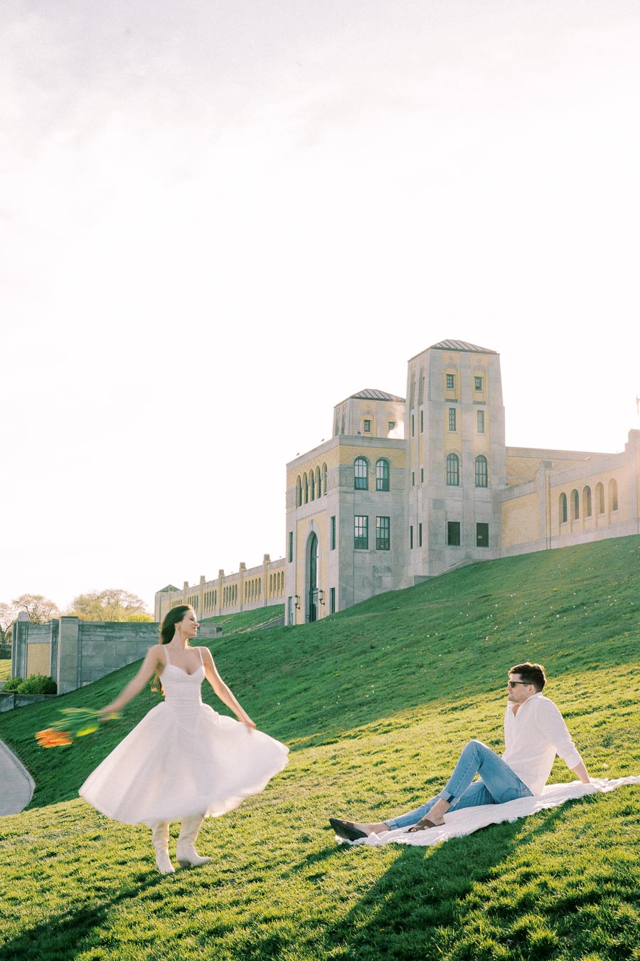 A woman in a white dress dances joyfully on a grassy hill next to a seated man, with a historic building in the background and a clear blue sky overhead.